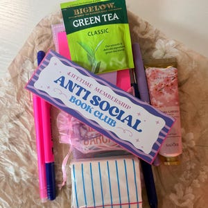 May include: A collection of items including a Bigelow Green Tea packet, a bookmark with "ANTI SOCIAL BOOK CLUB" text, pink pens, a small hand cream tube, and a small notepad with blue stripes. The items are arranged on a patterned surface.
