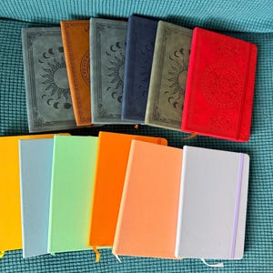 May include: A collection of eleven rectangular notebooks arranged in two rows. The top row features six notebooks with embossed sun and moon designs in grey, brown, dark grey, navy, olive, and red. The bottom row contains five solid-coloured notebooks.