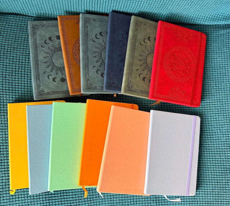 May include: A collection of eleven rectangular notebooks arranged in two rows. The top row features six notebooks with embossed sun and moon designs in grey, brown, dark grey, navy, olive, and red. The bottom row contains five solid-coloured notebooks.