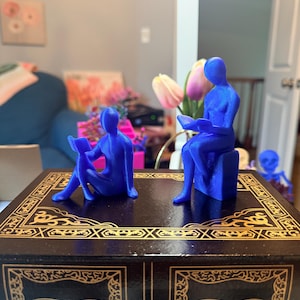 May include: Two blue 3D-printed figurines depicting abstract human forms reading books. One figure sits on the ground, while the other sits on a block. They are positioned on top of a dark brown rectangular box with gold decorative border patterns.