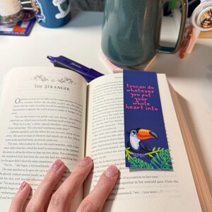 May include: A rectangular paper bookmark featuring a cartoon toucan perched on green leaves with the text "toucan do whatever you put your whole heart into" in pink, placed inside an open book with a dark green ceramic mug nearby.