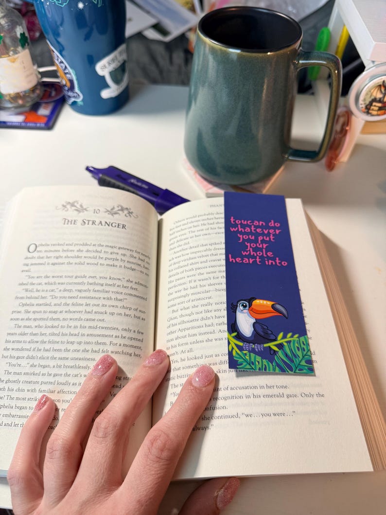 May include: A rectangular paper bookmark featuring a cartoon toucan perched on green leaves with the text "toucan do whatever you put your whole heart into" in pink, placed inside an open book with a dark green ceramic mug nearby.