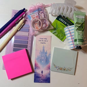 May include: A collection of stationery and personal care items including three pens, a set of purple sticky tabs, a pink organza bag with washi tape, a translucent hair claw, a green tea bag, a tube of hand cream, and two sets of sticky notes.