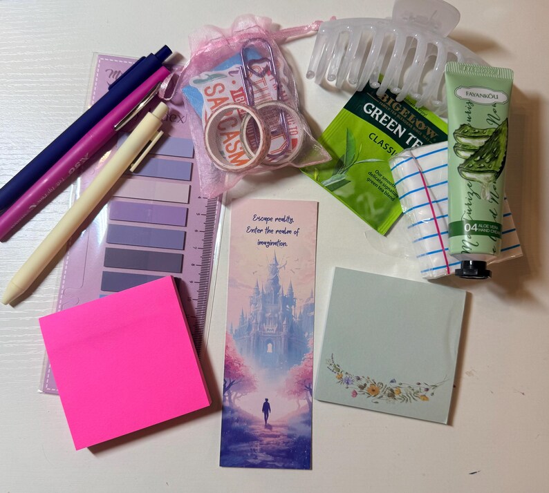 May include: A collection of stationery and personal care items including three pens, a set of purple sticky tabs, a pink organza bag with washi tape, a translucent hair claw, a green tea bag, a tube of hand cream, and two sets of sticky notes.
