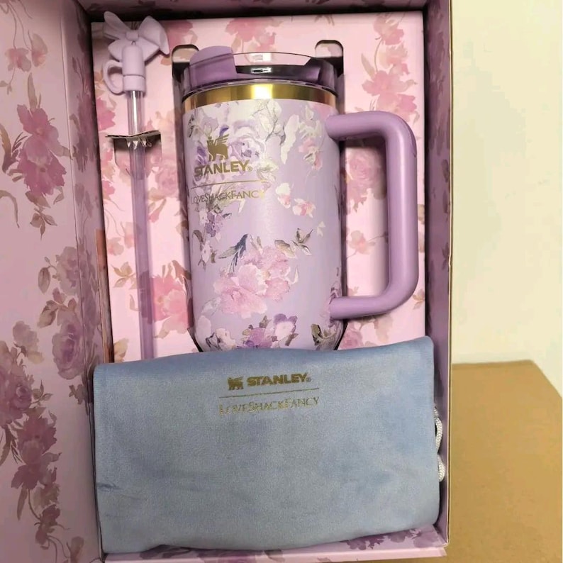 K&ouml;nnte beinhalten: Ein lavendelfarbener Stanley-Becher mit Blumenmuster, passendem Strohhalm mit Schleifenaufsatz und einem hellblauen Beutel, alles in einer Geschenkbox mit Blumenmuster. Der Becher tr&auml;gt die Logos Stanley und LoveShackFancy in Gold.