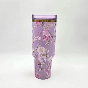 K&ouml;nnte beinhalten: Ein hoher, zylindrischer Edelstahlbecher in einem sanften Lavendelton mit einem detaillierten Blumenmuster in verschiedenen Rosa-, Lila- und Wei&szlig;t&ouml;nen. Der Becher verf&uuml;gt &uuml;ber einen metallisch goldenen Rand und einen passenden, transparenten Kunststoffdeckel.