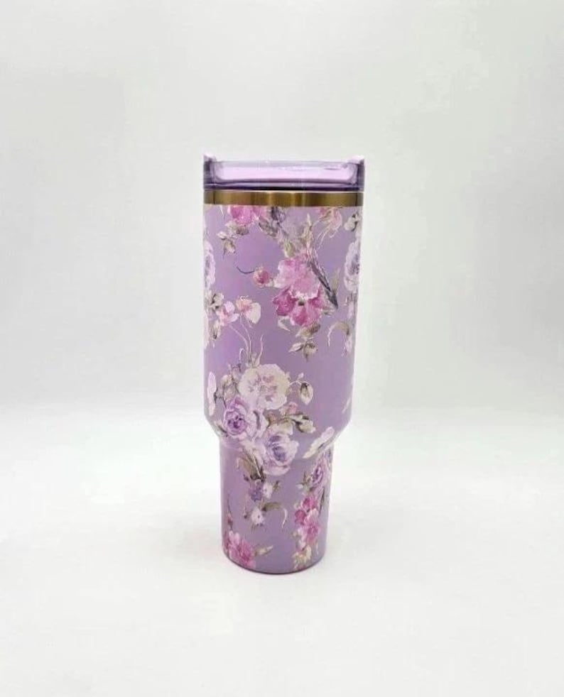 K&ouml;nnte beinhalten: Ein hoher, zylindrischer Edelstahlbecher in einem sanften Lavendelton mit einem detaillierten Blumenmuster in verschiedenen Rosa-, Lila- und Wei&szlig;t&ouml;nen. Der Becher verf&uuml;gt &uuml;ber einen metallisch goldenen Rand und einen passenden, transparenten Kunststoffdeckel.