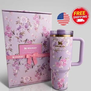 K&ouml;nnte beinhalten: Ein lavendelfarbener Stanley-Becher mit Blumenmuster und passender Geschenkbox. Der Becher hat einen goldenen Rand, einen violetten Henkel und einen transparenten Deckel. Die Box ist mit demselben Blumenmuster, einer rosa Schleife und dem Stanley LoveShackFancy-Logo versehen.