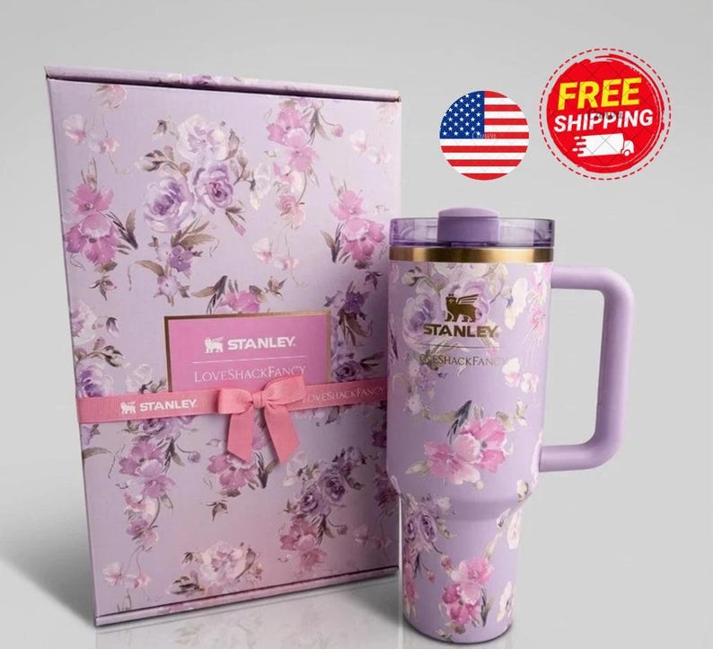 K&ouml;nnte beinhalten: Ein lavendelfarbener Stanley-Becher mit Blumenmuster und passender Geschenkbox. Der Becher hat einen goldenen Rand, einen violetten Henkel und einen transparenten Deckel. Die Box ist mit demselben Blumenmuster, einer rosa Schleife und dem Stanley LoveShackFancy-Logo versehen.