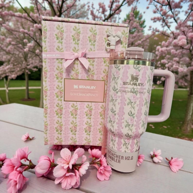 May include: A Stanley Quencher H2.0 tumbler and its matching box, both featuring a pink and white floral pattern with vertical stripes. The tumbler has a light pink handle, a clear lid, and a straw. The box is tied with a pink ribbon.