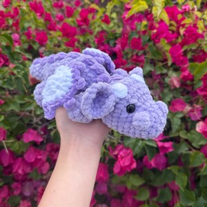 May include: A handmade, plush dragon toy in shades of lavender and white. The dragon has a textured, crocheted appearance with a black eye. The wings and belly are white, while the rest of the dragon is lavender.