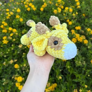 May include: A handmade crochet giraffe plush toy in yellow and brown. The giraffe has black eyes and a white muzzle. The plush is held against a background of yellow flowers and green foliage.