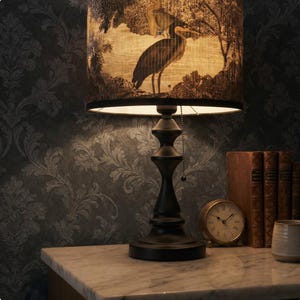 May include: A black table lamp with a heron design lampshade. The lampshade features a black and white illustration of two herons in a natural setting. The lamp has a dark, ornate base and sits on a marble surface.