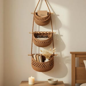 Puede incluir: Tres cestas colgantes de almacenamiento de hilo de crochet marrón. Las cestas están suspendidas de una cuerda y tacos de madera. La cesta superior contiene un libro con la palabra "Home". La cesta inferior contiene libros y un jarrón pequeño.