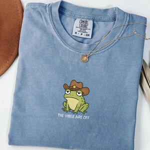 Peut inclure: Un t-shirt bleu à manches courtes avec une broderie représentant une grenouille verte portant un chapeau de cowboy marron avec une étoile jaune. Sous la grenouille, le texte indique THE VIBES ARE OFF. Étiquette Comfort Colors au col.