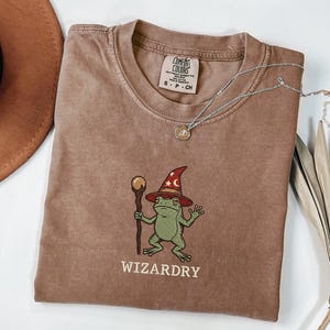 Può includere: Una t-shirt marrone a maniche corte con la grafica di una rana verde che indossa un cappello da mago rosso con stelle e tiene in mano un bastone di legno con una sfera dorata. Il testo WIZARDRY è stampato sotto la rana. Etichetta Comfort Colors.