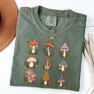 May include: A sage green short-sleeve t-shirt featuring a graphic print of nine different mushrooms in various colors including red, brown, purple, blue, and pink, accented with small leaves and star shapes. The shirt has a Comfort Colors brand label.