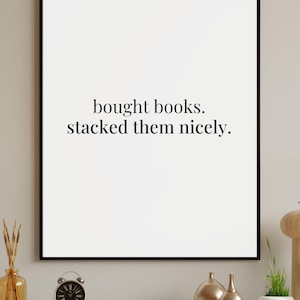 Può includere: Stampa incorniciata con il testo "bought books. stacked them nicely." in nero. Sotto, una mensola con una piccola sveglia, vasi dorati e altri oggetti decorativi.