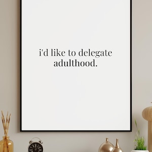 May include: A framed print with the text "I'd like to delegate adulthood." The print is in a black frame and is displayed above a shelf with decorative items, including a small alarm clock and vases.