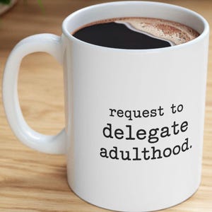 May include: A white ceramic mug filled with dark coffee, featuring a handle on the left side. The front of the mug displays the black printed text "request to delegate adulthood." in a simple, sans-serif font, centered on the smooth surface.