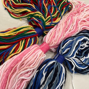 May include: Close-up of several colorful yarn tassels. The tassels are in various colors, including red, yellow, green, blue, and pink. Each tassel is made of multiple strands of yarn tied together at the top with a fabric band.