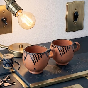Peut inclure: Deux tasses en terre cuite de forme arrondie avec une anse, ornées d'un motif peint à la main en noir composé de petites tirets verticaux autour du bord supérieur. Les tasses sont posées sur une surface sombre près d'une bobine de fil bleu et or.