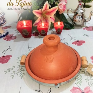 Peut inclure: Un plat à tajine en terre cuite avec un bord en métal et des poignées en bois est posé sur une nappe à motifs floraux. Trois bougies rouges allumées et une théière en argent sont en arrière-plan. Le texte "L'Artisanat du Tajine Authenticity & Saveur" est en haut.