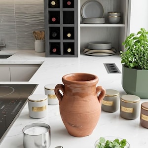 Peut inclure: Une jarre en terre cuite de style amphore est posée sur un comptoir en marbre blanc, entourée de plusieurs petits pots à épices cylindriques avec des couvercles dorés étiquetés Saffron et Cumin, un bol argenté et une plante aromatique verte.
