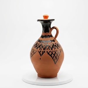 May include: A terracotta-colored ceramic jug with a black and orange lid and handle. The jug features a black patterned design around the neck and body. The jug is displayed on a white surface.
