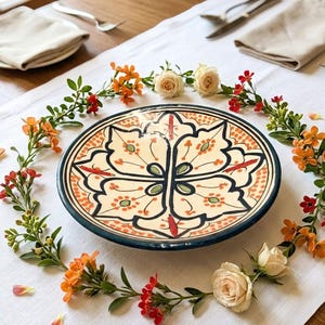 Peut inclure: Une assiette décorative en céramique avec une base crème et un motif floral détaillé en orange, vert et noir. L'assiette est entourée d'une guirlande de fleurs orange, de fleurs rouges et de roses blanches, posée sur une nappe blanche.