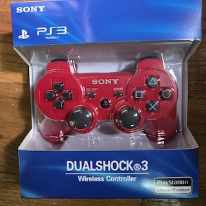 May include: A red DualShock 3 wireless controller for the PlayStation 3 console, packaged in a blue and white box. The controller has black buttons and the Sony logo. The box also displays the PlayStation logo and the words "Official Product."