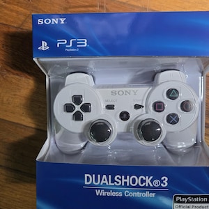 May include: A white DualShock 3 wireless controller for PlayStation 3, packaged in a blue and white box. The controller has black analog sticks and buttons with colored symbols. The box features the Sony and PlayStation logos.