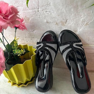 May include: A pair of black slip-on sandals featuring a glossy finish, a rounded toe, and a wide strap across the top. The strap is constructed from a sheer mesh material overlaid with a decorative pattern of small, dark, reflective rhinestones.