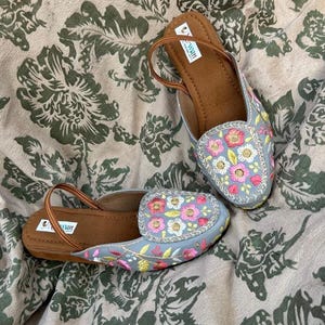 Puede incluir: Un par de bailarinas bordadas en azul claro con diseños florales en rosa, amarillo y blanco. Los zapatos tienen plantillas y correas marrones. El fondo es una tela con estampado floral.