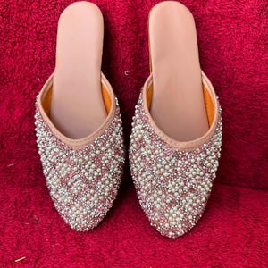May include: A pair of slip-on mules featuring a pointed toe design. The exterior is densely embellished with small white faux pearls and shimmering silver-toned crystals arranged in a textured pattern. The interior lining is a smooth, light beige material.