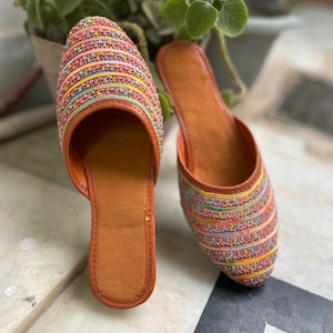 Könnte beinhalten: Ein Paar Slipper mit abgerundeter Zehenpartie und flacher Sohle. Der obere Teil des Schuhwerks ist mit horizontalen Reihen aus mehrfarbigen Stickereien in Rosa, Gelb, Blau und Grün verziert und mit einer hellbraunen Einfassung versehen.