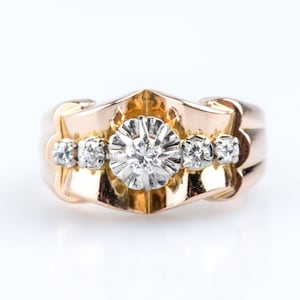 May include: A gold-toned ring with a central diamond and four smaller diamonds. The ring has a decorative, geometric design with a bright, polished finish. The ring is set against a white background.
