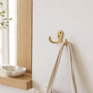 Könnte beinhalten: Ein doppelter Wandhaken aus Messing, der an einer weißen Oberfläche montiert ist und ein geschwungenes Design mit zwei sichtbaren Schraublöchern zur Befestigung aufweist. Ein beigefarbener Stofftaschenriemen hängt am unteren Haken.