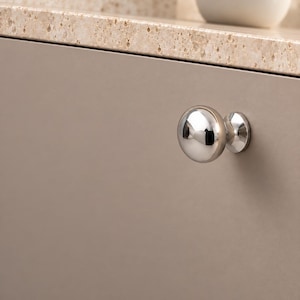 May include: A polished chrome cabinet knob with a smooth, rounded, mushroom-shaped head and a circular base, mounted on a flat, solid taupe-colored cabinet door panel. The metallic surface reflects the surrounding light and environment.