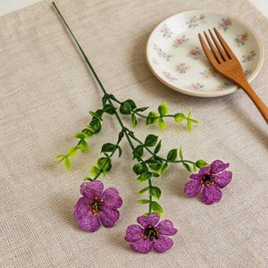 May include: A sprig of artificial flowers with three purple blossoms and green leaves. A small floral-patterned plate and a wooden fork are also present on a beige linen surface.
