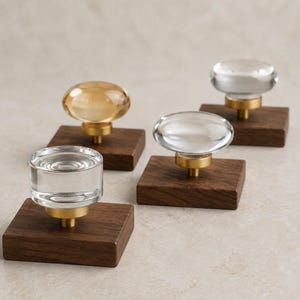 May include: Four cabinet knobs, each featuring a clear or amber-tinted glass top mounted on a circular brass-toned base, which is attached to a square dark brown wooden block. The knobs vary in shape between rounded and cylindrical glass forms.