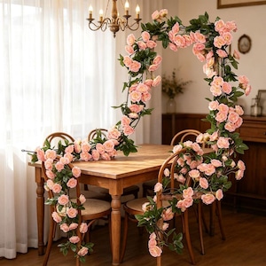 Puede incluir: Un arco decorativo construido con enredaderas artificiales que presenta numerosas flores de rosas de color rosa suave y hojas de color verde oscuro. La guirnalda floral está colocada sobre una mesa de comedor de madera y sillas.