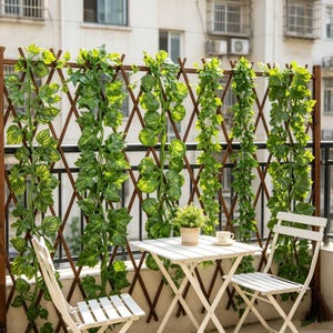 Puede incluir: Una valla de celosía extensible de madera marrón decorada con hileras verticales de hojas de hiedra artificiales verdes. La valla está situada detrás de una mesa plegable blanca y dos sillas plegables blancas, con una pequeña planta en maceta.