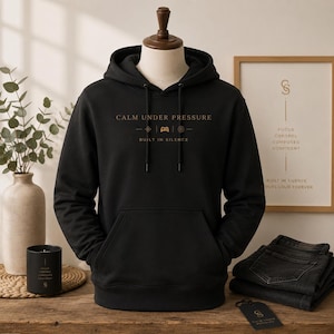 May include: Black hoodie with the text "CALM UNDER PRESSURE" in gold lettering. The hoodie has a front pocket and a drawstring hood. The words "BUILT IN SILENCE" are below the main text. A framed print and a candle are in the background.