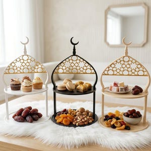Könnte beinhalten: Dreistöckige Servierstände in Weiß, Schwarz und Beige, jeweils mit Moschee-Design. Sie präsentieren Cupcakes, Macarons, Datteln und Nüsse. Die Ständer haben ein goldenes geometrisches Muster und einen Halbmond. Ideal zum Servieren von Leckereien.