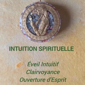 May include: A circular pendant necklace with a gold-colored Egyptian figure and hieroglyphs. The pendant is filled with small, colorful stones and hangs from a black cord. The text "INTUITION SPIRITUELLE" is displayed below the pendant.