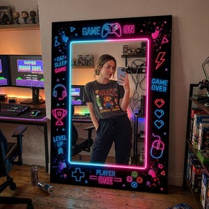 Può includere: Un grande specchio con cornice nera e bordo blu e rosa neon. Lo specchio presenta grafica e testo a tema gaming, tra cui "Game On", "Game Over" e "Player One". Lo specchio si trova in una stanza con attrezzature di gioco.