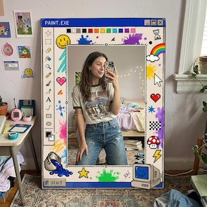 Pode incluir: Um espelho retangular com uma moldura azul projetada para se parecer com uma janela de computador, com o texto "PAINT.EXE" no topo. O espelho é decorado com respingos de tinta coloridos, ícones pixelados e um arco-íris. O espelho é um item decorativo.