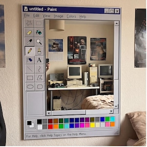 Può includere: Uno specchio da parete progettato per assomigliare all'interfaccia della classica applicazione Windows Paint. La cornice è grigia con una barra superiore blu e una tavolozza di colori in basso. Lo specchio riflette una stanza con computer e poster vintage. Il testo "untitled - Paint" è visibile.