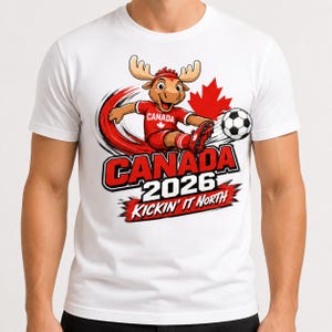 May include: A white t-shirt featuring a cartoon moose wearing a red soccer uniform and kicking a soccer ball. The graphic includes a red maple leaf, the text CANADA 2026, and the phrase KICKIN' IT NORTH in bold red and black lettering.
